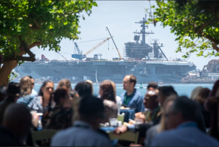 Attendees at the Government Contract Pricing Summit in San Diego, with a naval carrier in the bay behind them